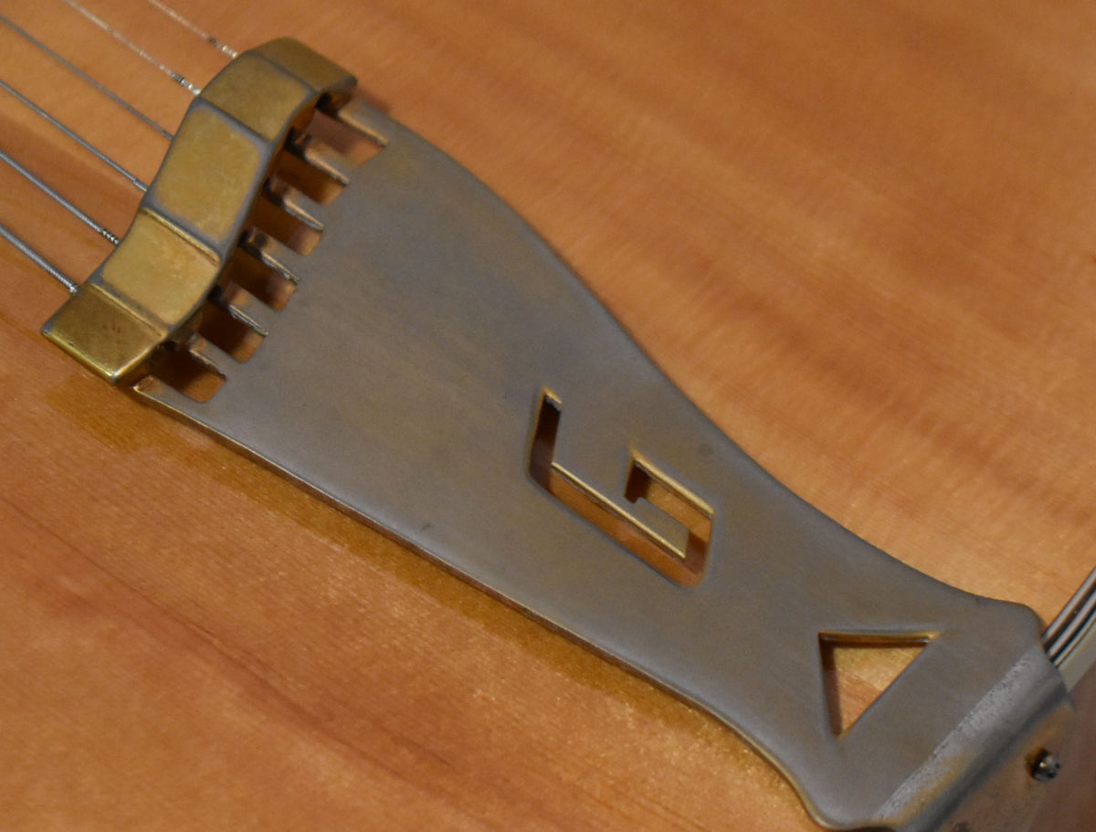Close-up of a guitar's tailpiece, showing a brushed metal plate with a cutout "G" logo and a brass string holder.