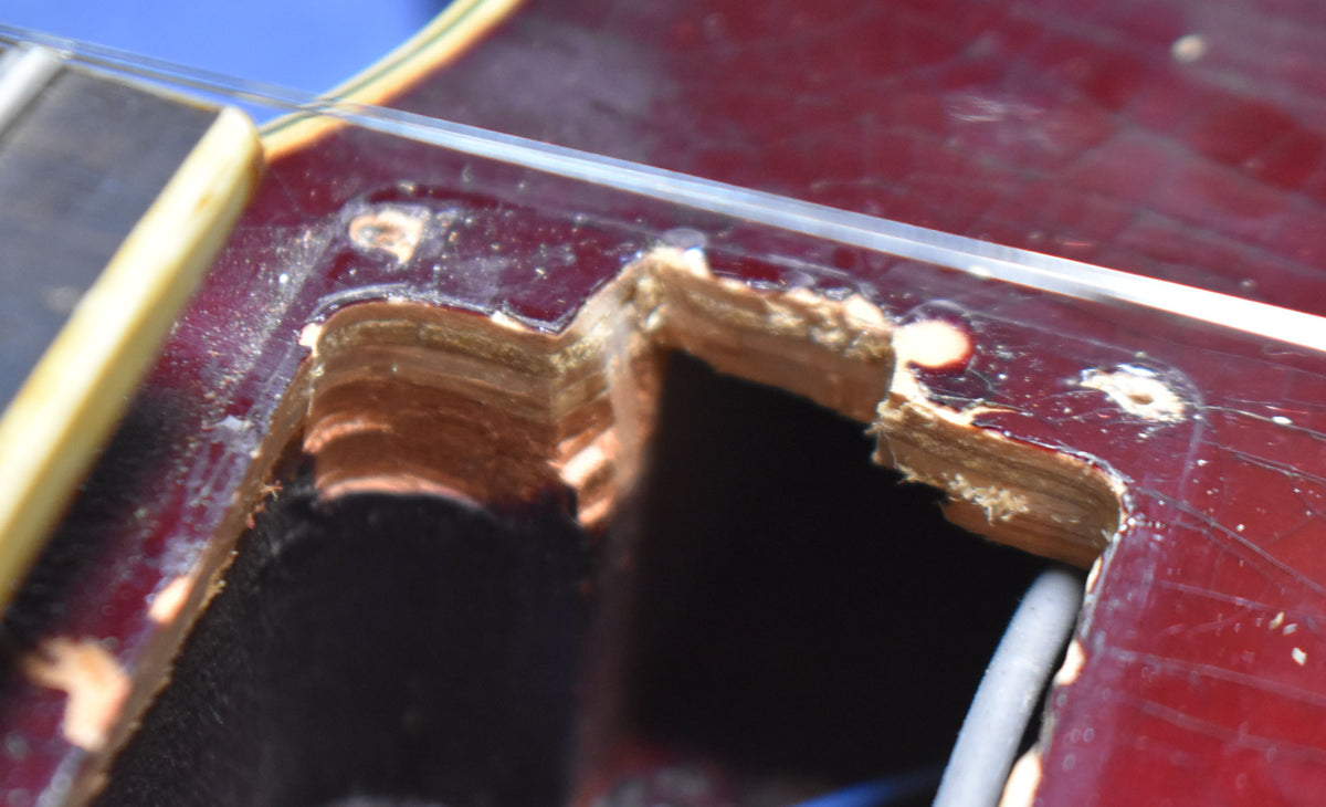 Close-up of the guitar's dark red body, showing a chipped-out control cavity with exposed wood layers and a wire emerging from the dark interior.
