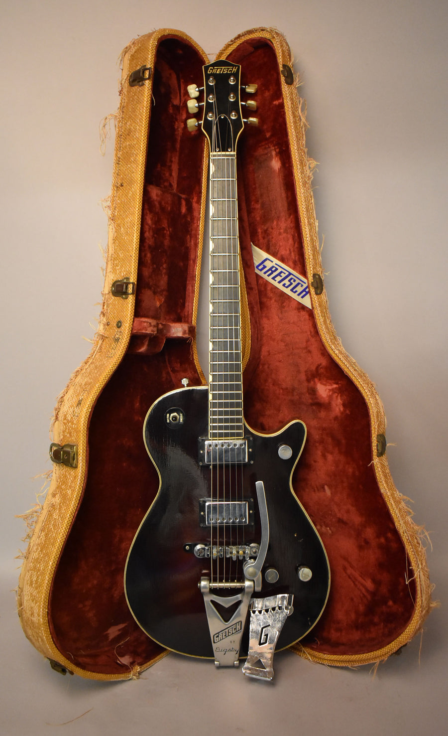 Full-length view of a black electric guitar with a Bigsby tremolo, gold hardware, and white binding, resting in its open, worn, red-lined case.