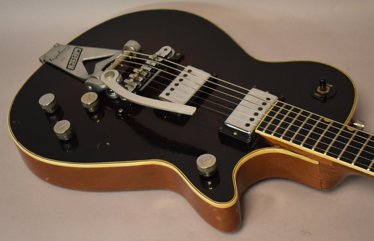 Close-up of a black electric guitar with a Bigsby vibrato, two humbuckers, four chrome knobs, and a three-way switch.