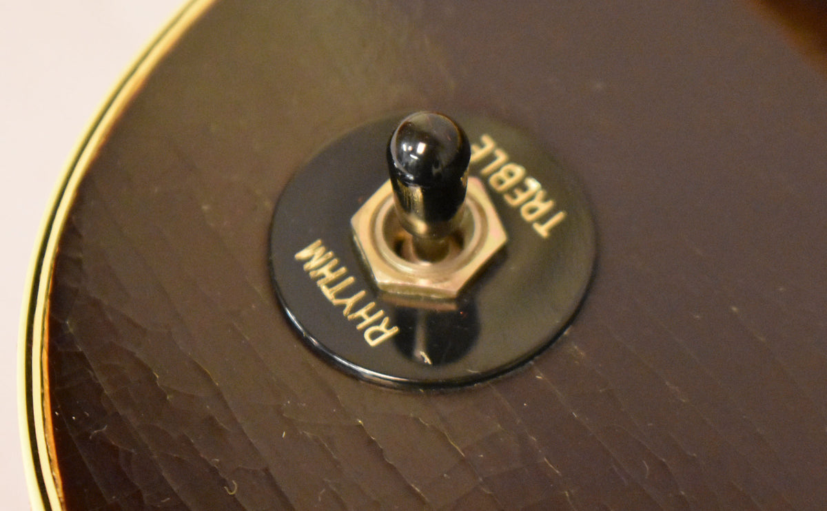 Close-up of the guitar's dark brown body, showing a black pickup selector switch with "RHYTHM" and "TREBLE" in gold lettering.