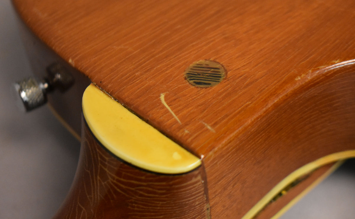 Close-up of the guitar's neck heel and lower bout, showing a natural wood finish with checking, a yellow binding, and a circular sticker.