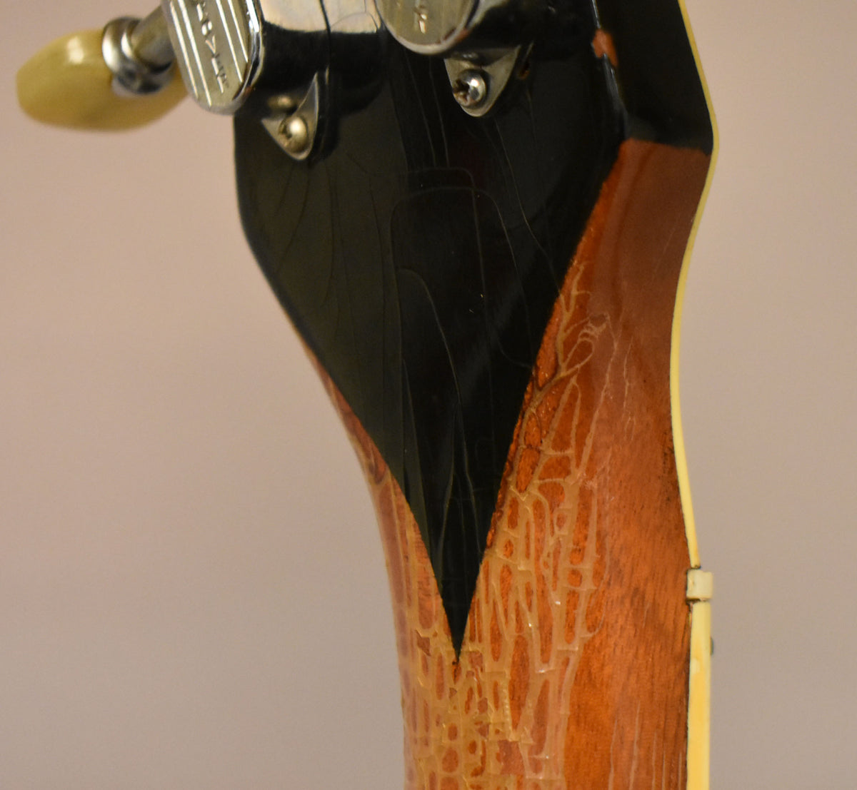Close-up of the guitar's headstock and neck heel, showing heavy finish checking on the black headstock and amber-stained neck.