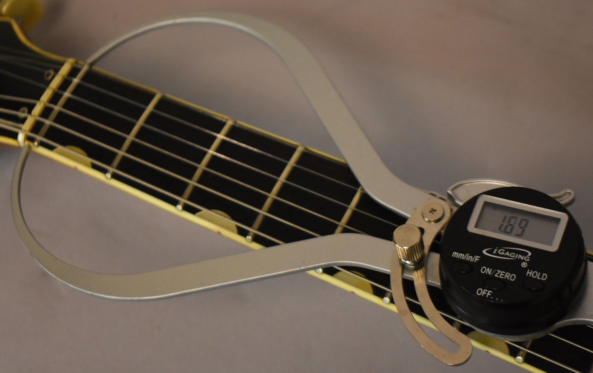 A digital caliper measures the nut width of the guitar's black fretboard with cream binding, showing 1.69 on the display.