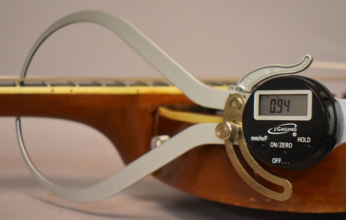 A digital caliper measures the neck thickness at the first fret, showing 0.94 inches, with the fretboard and binding visible.