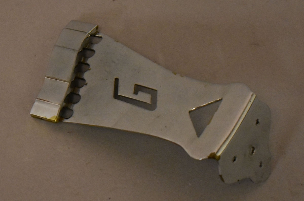 Close-up of the chrome tailpiece, showing the "G" cutout, string bar, and mounting holes, with some tarnishing on the edges.
