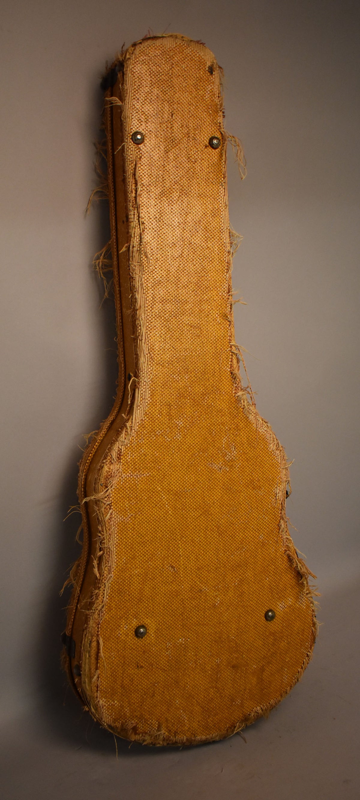 The guitar's original hard shell case, covered in heavily frayed, textured tan fabric, stands upright against a gray background.