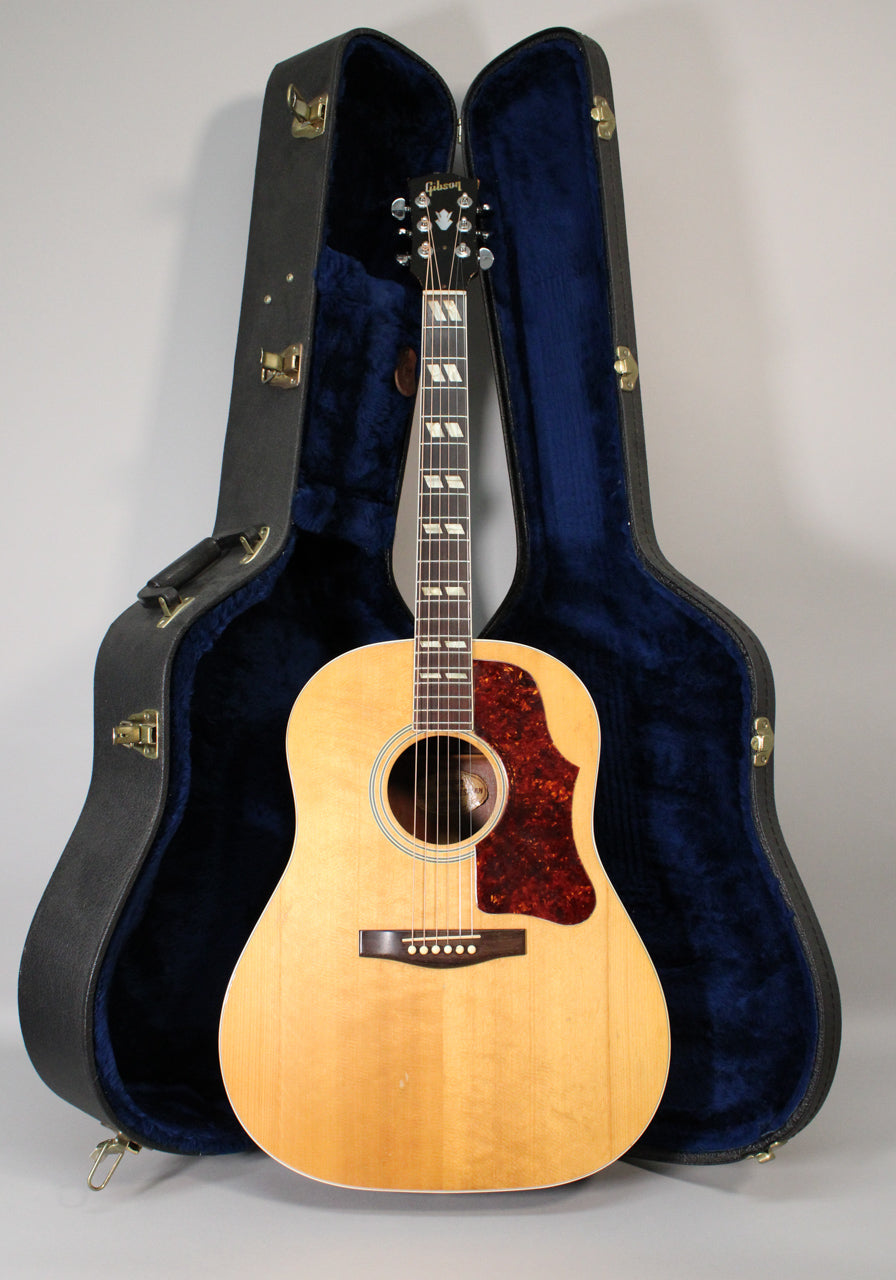 Full-front view of a natural finish acoustic guitar with a dark tortoiseshell pickguard, standing in its open, blue-lined hard case.