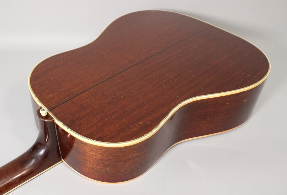 The guitar's dark brown back and sides, with cream binding, show light scratches, finish wear, and a visible center seam.