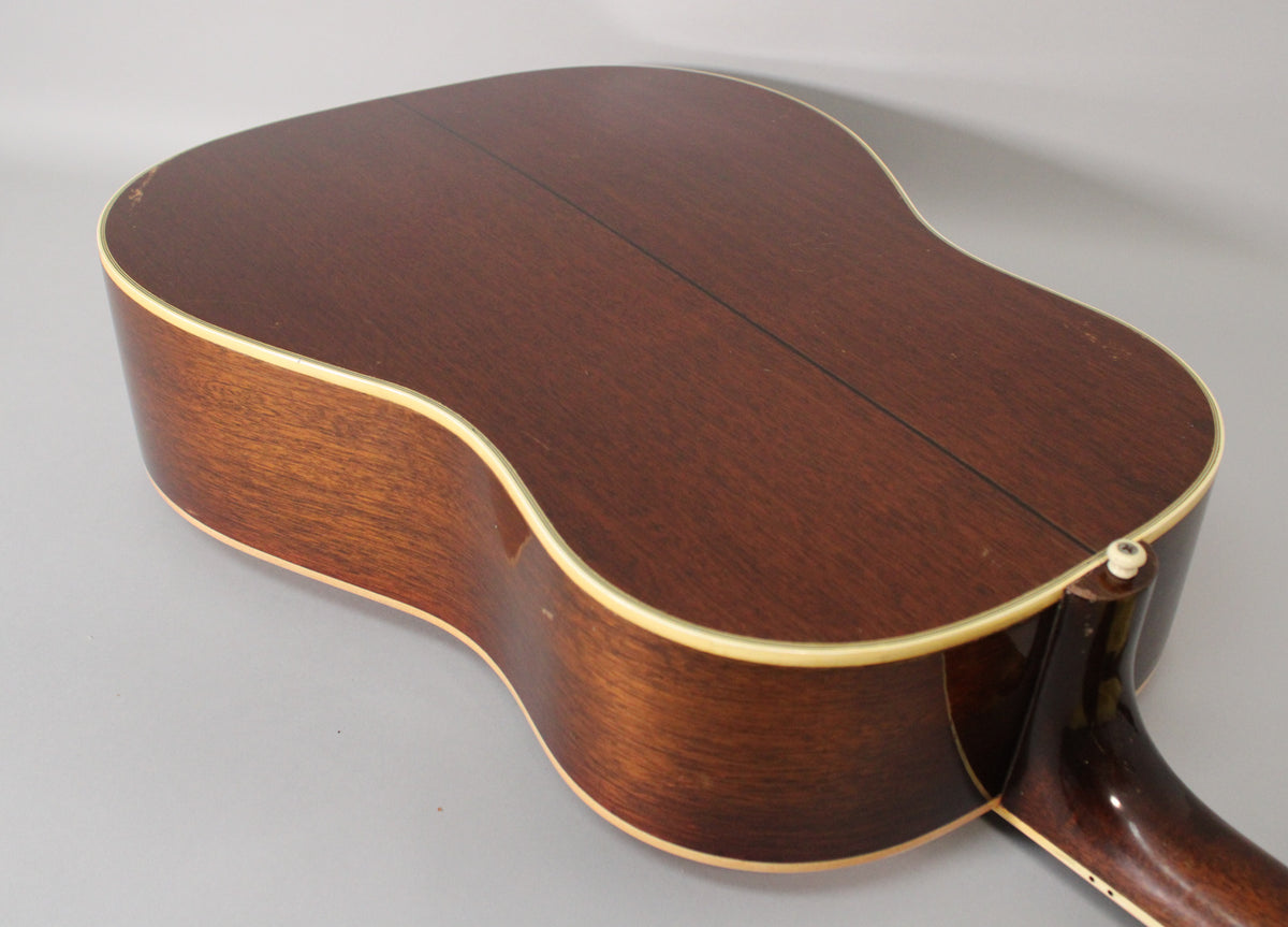 The acoustic guitar's dark brown back and sides are angled, revealing a center seam, cream binding, and a few chips on the upper bout.