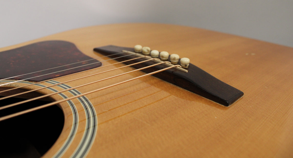 Close-up of the natural finish acoustic guitar's body, showing the dark pickguard, bridge with six ivory-colored pins, and soundhole rosette.