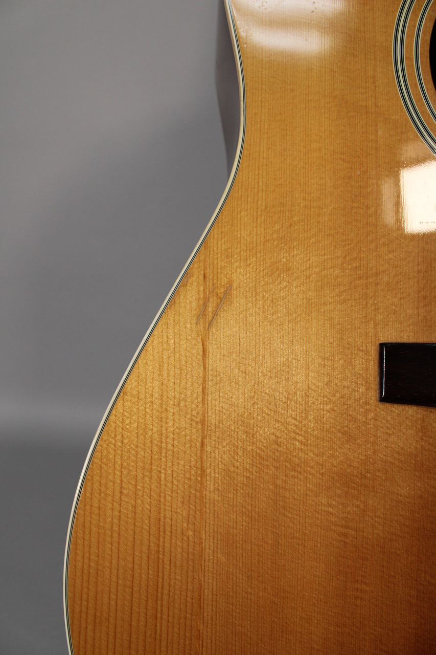 Close-up of the natural-finish acoustic guitar's lower bout, showing two diagonal scratches on the upper left and a small part of the bridge.