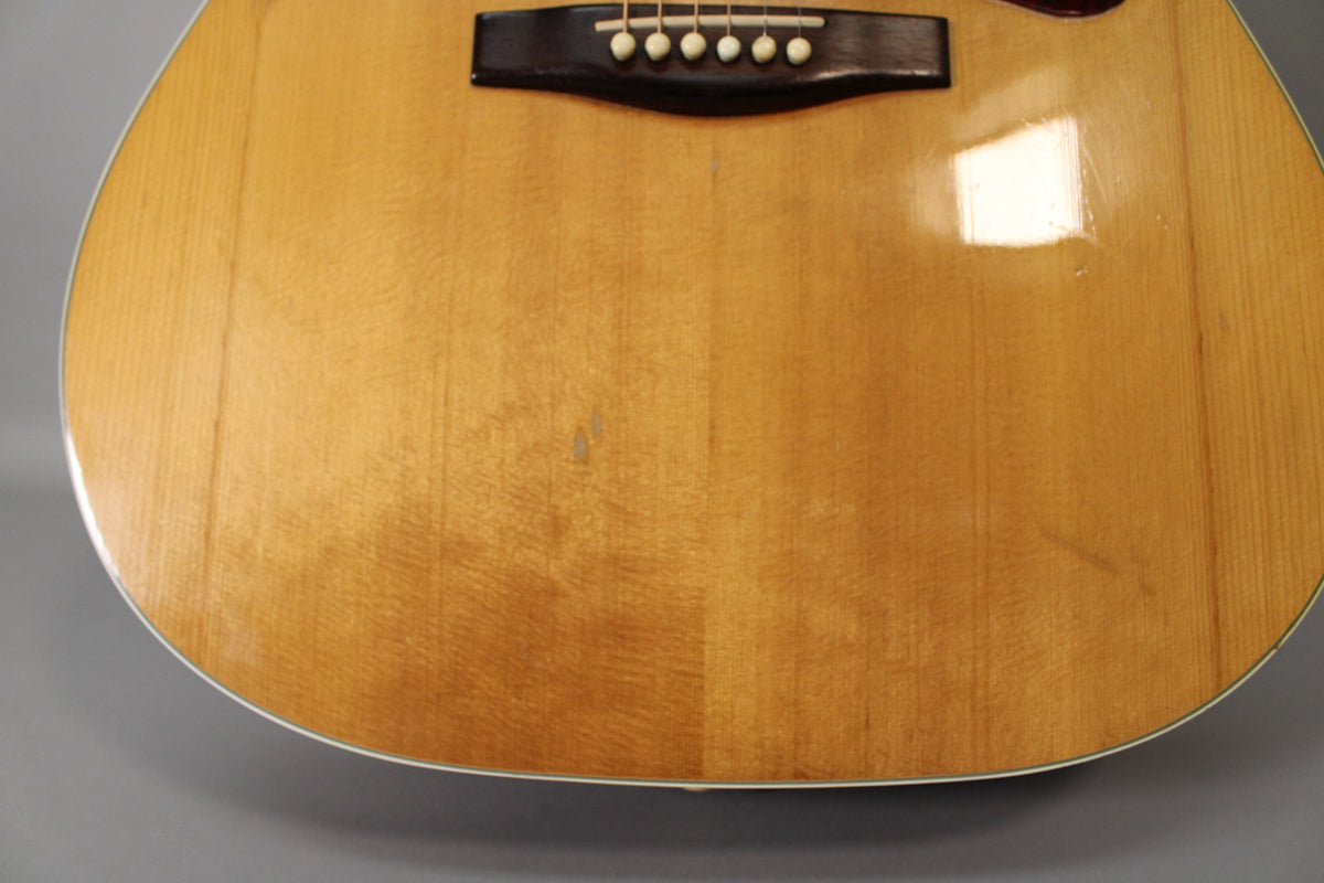The lower half of the natural finish acoustic guitar body, showing the dark bridge with six white pins and some light play wear.