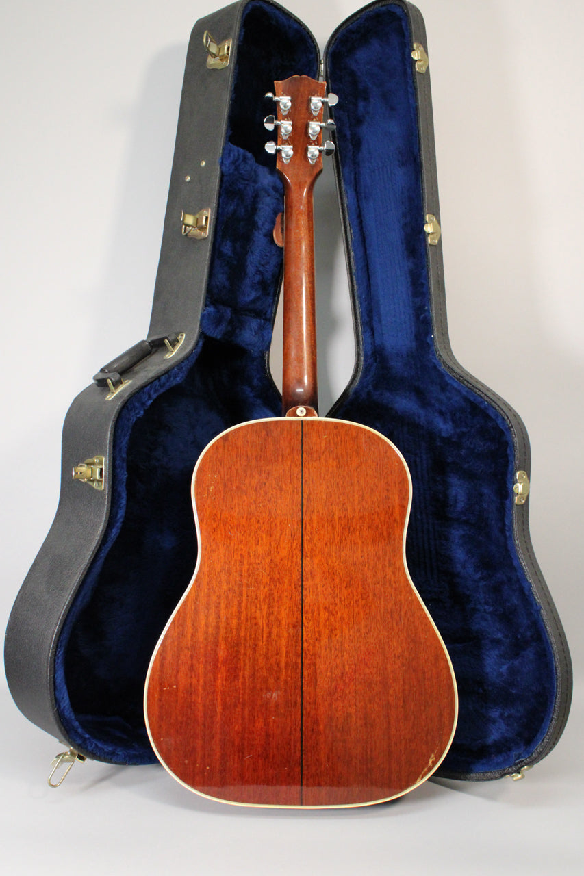 The guitar's natural finish back and neck are visible, with some wear on the lower bout, standing in an open hard case.