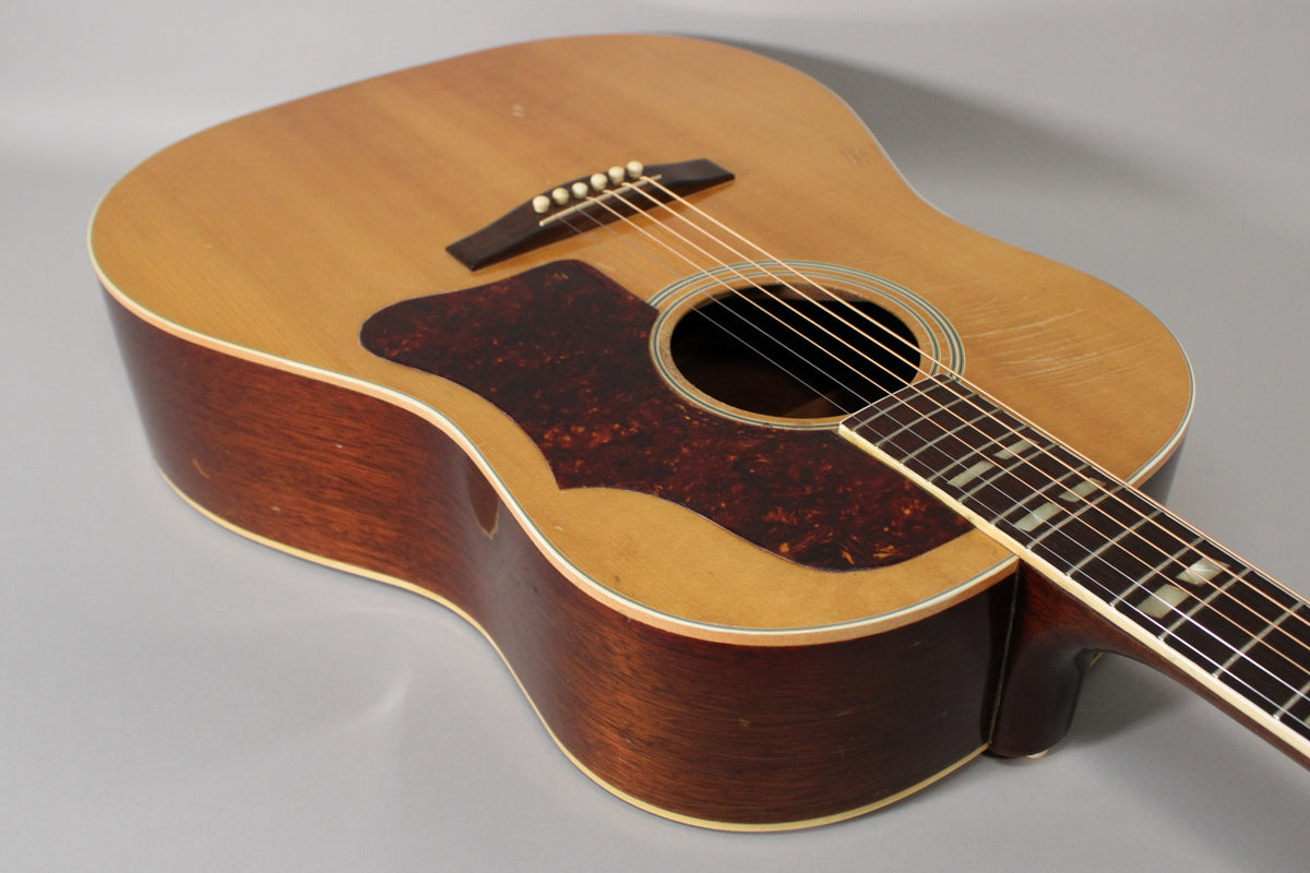 The natural finish acoustic guitar is angled from the upper left, showing its light-colored top, dark pickguard, and mahogany sides.