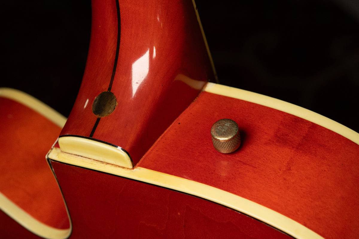 Close-up of the guitar's orange body and neck joint, showing a strap button, a missing strap button, and cream binding.