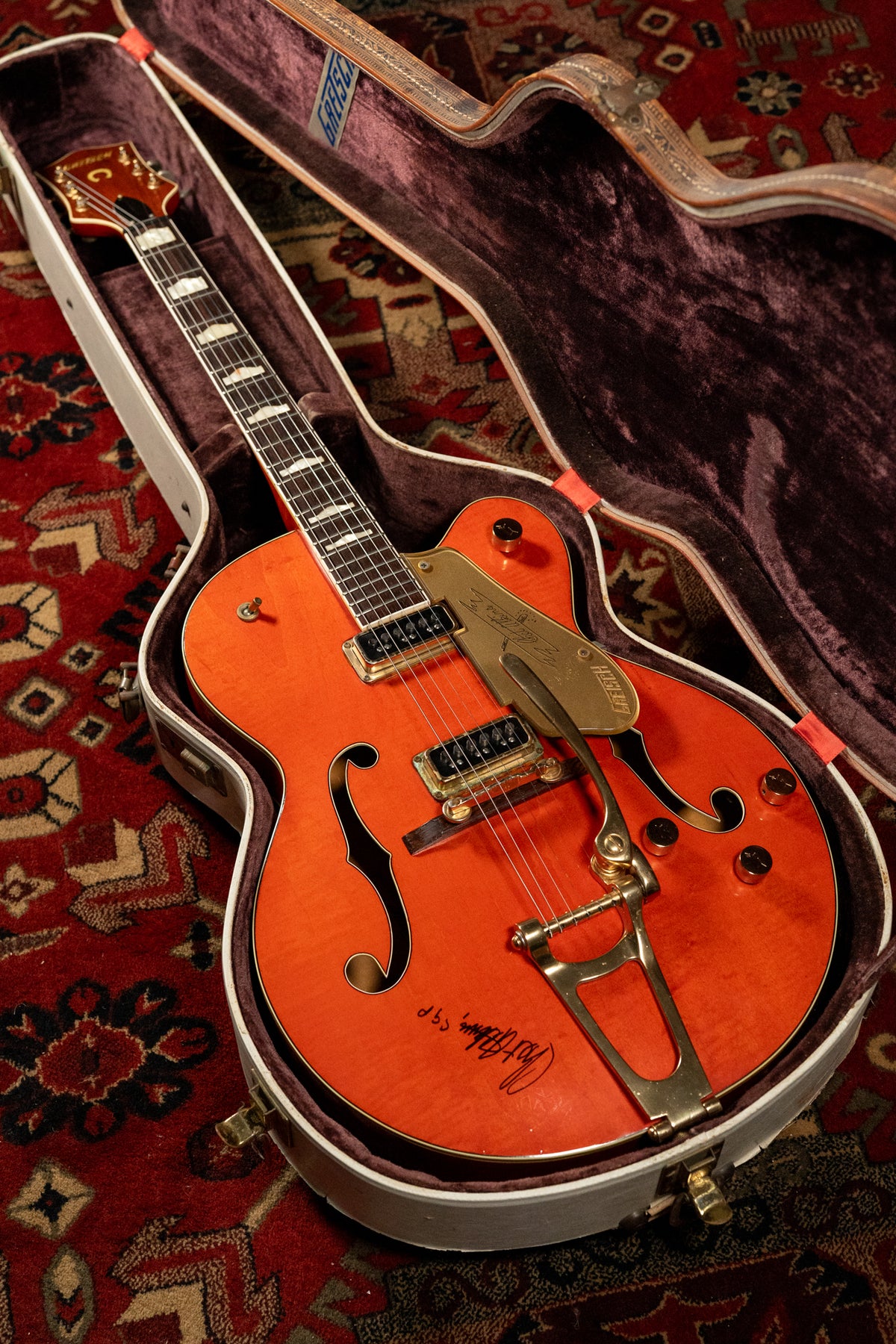 Orange hollow-body electric guitar in a plush purple-lined case, with gold hardware, f-holes, and a visible signature on the body.