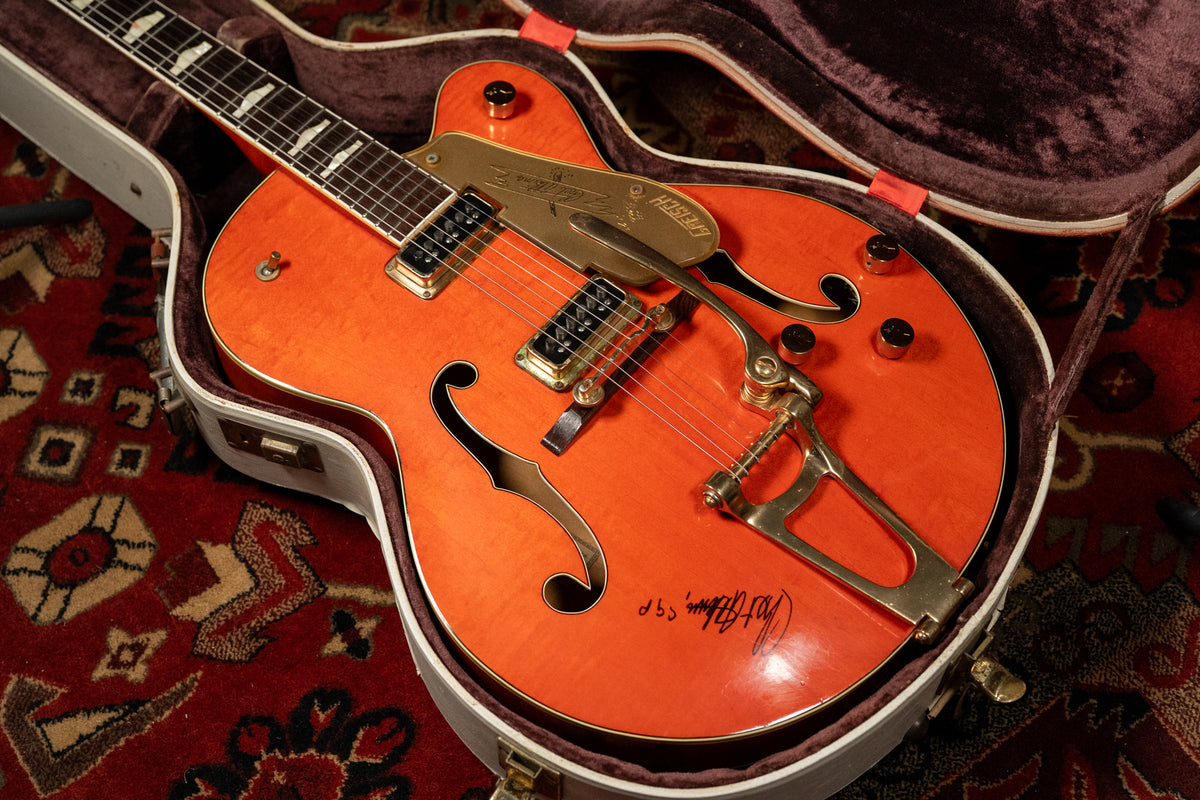 The orange hollow-body guitar, with gold hardware and f-holes, rests in its open case, showing a signature on the lower bout.