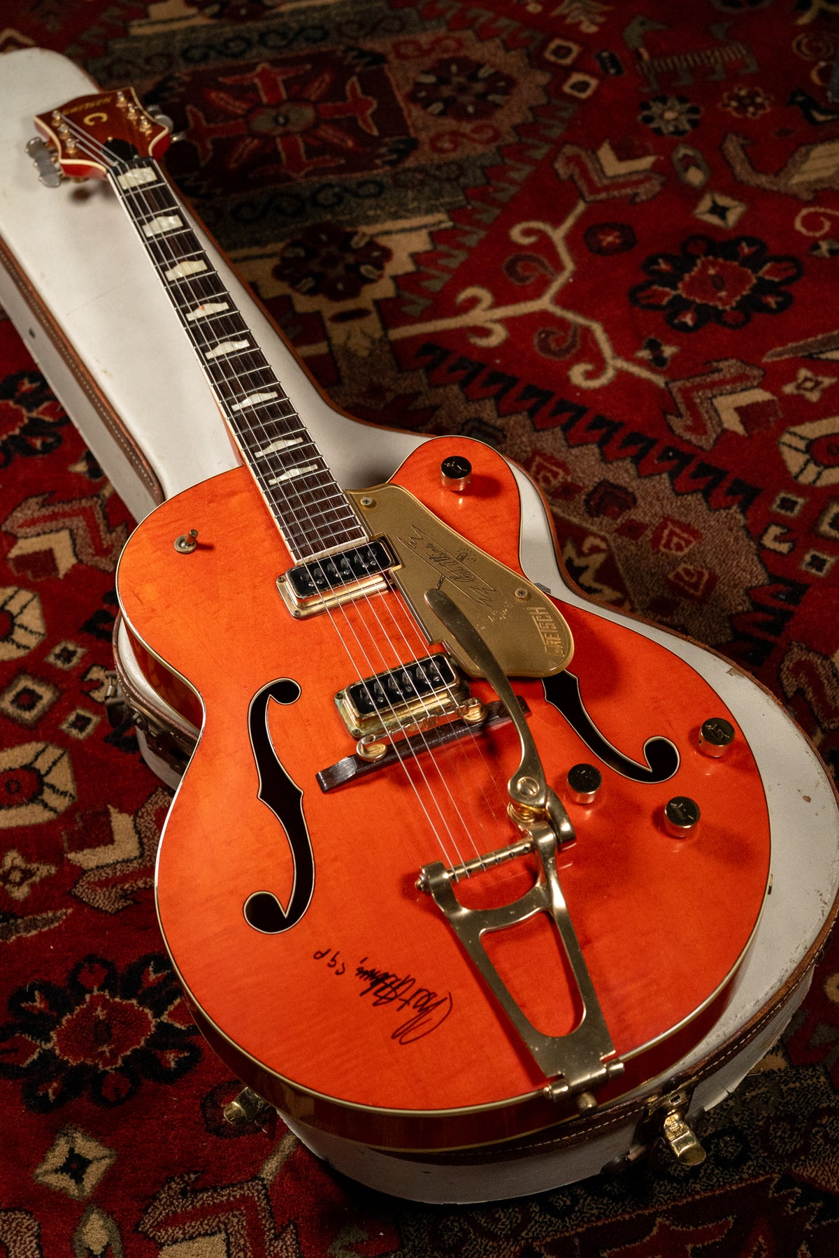 Orange hollow-body electric guitar with gold hardware, f-holes, and a Bigsby tremolo, resting in a white case.