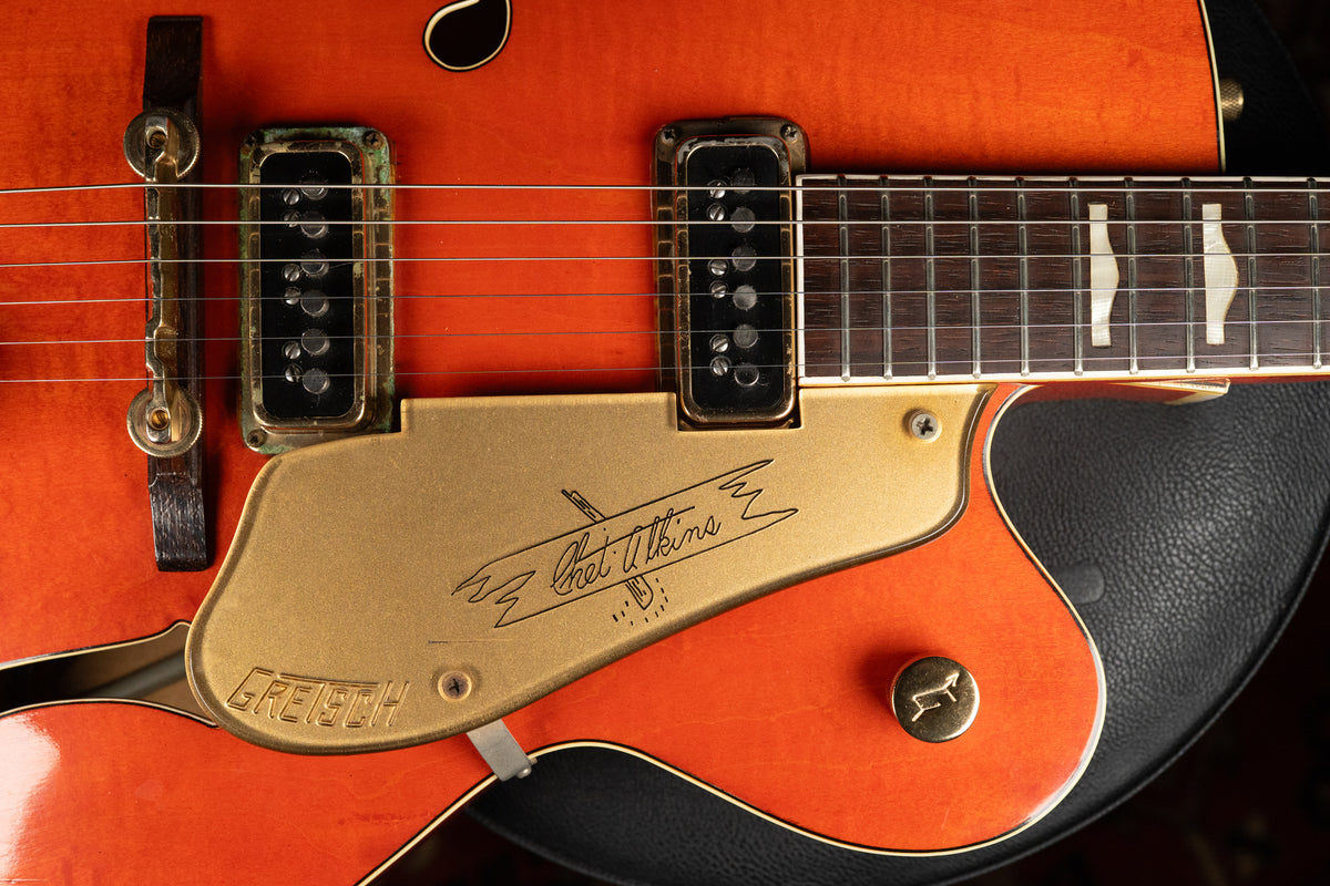 Close-up of the orange guitar body, showing the gold pickguard with signature, neck pickup with green corrosion, and a control knob.