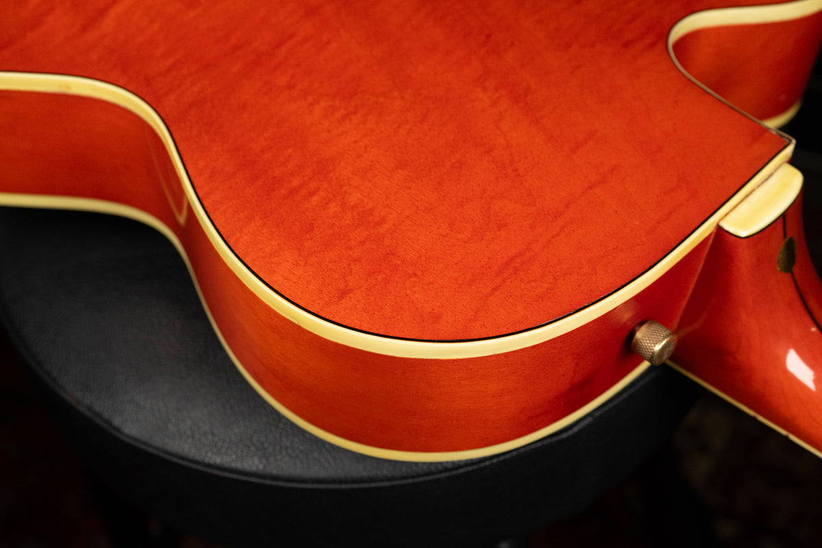 Close-up of the guitar's lower bout, showing the orange finish with wood grain, cream binding, and a brass strap button.