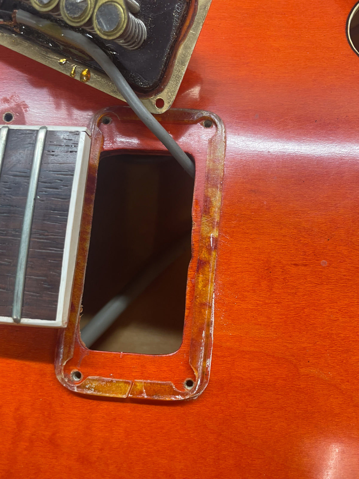Close-up of the orange guitar body, showing a rectangular control cavity with a clear plastic surround and a pickup removed.