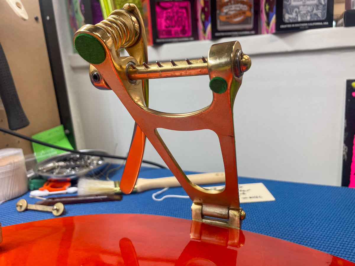 Close-up of the guitar's gold Bigsby vibrato tailpiece, showing its arm, spring, and mounting to the glossy orange body.