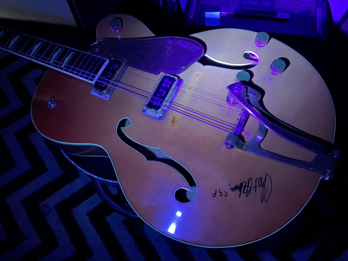 Orange hollow-body electric guitar, angled from neck to body, showing f-holes, Bigsby vibrato, and a signature on the lower bout.
