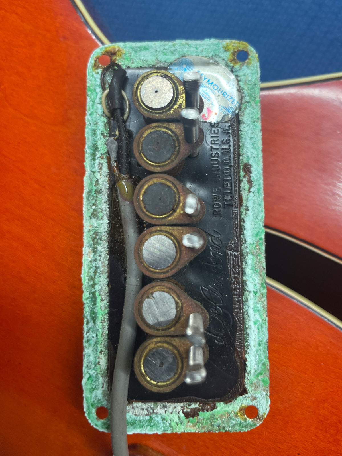 Close-up of a pickup's underside, showing six rusty pole pieces, wiring, and a corroded green felt pad on an orange guitar body.