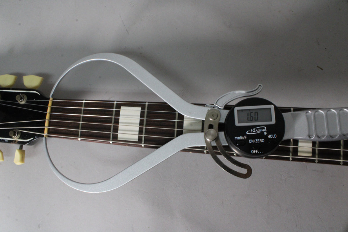 A digital caliper measures the guitar's dark fretboard with rectangular inlays, showing 1.60mm, near the headstock with cream tuners.
