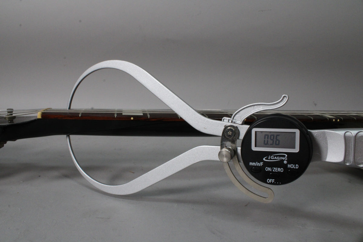 A digital caliper measures the neck relief of the guitar's dark fretboard, displaying "0.96" on its screen.