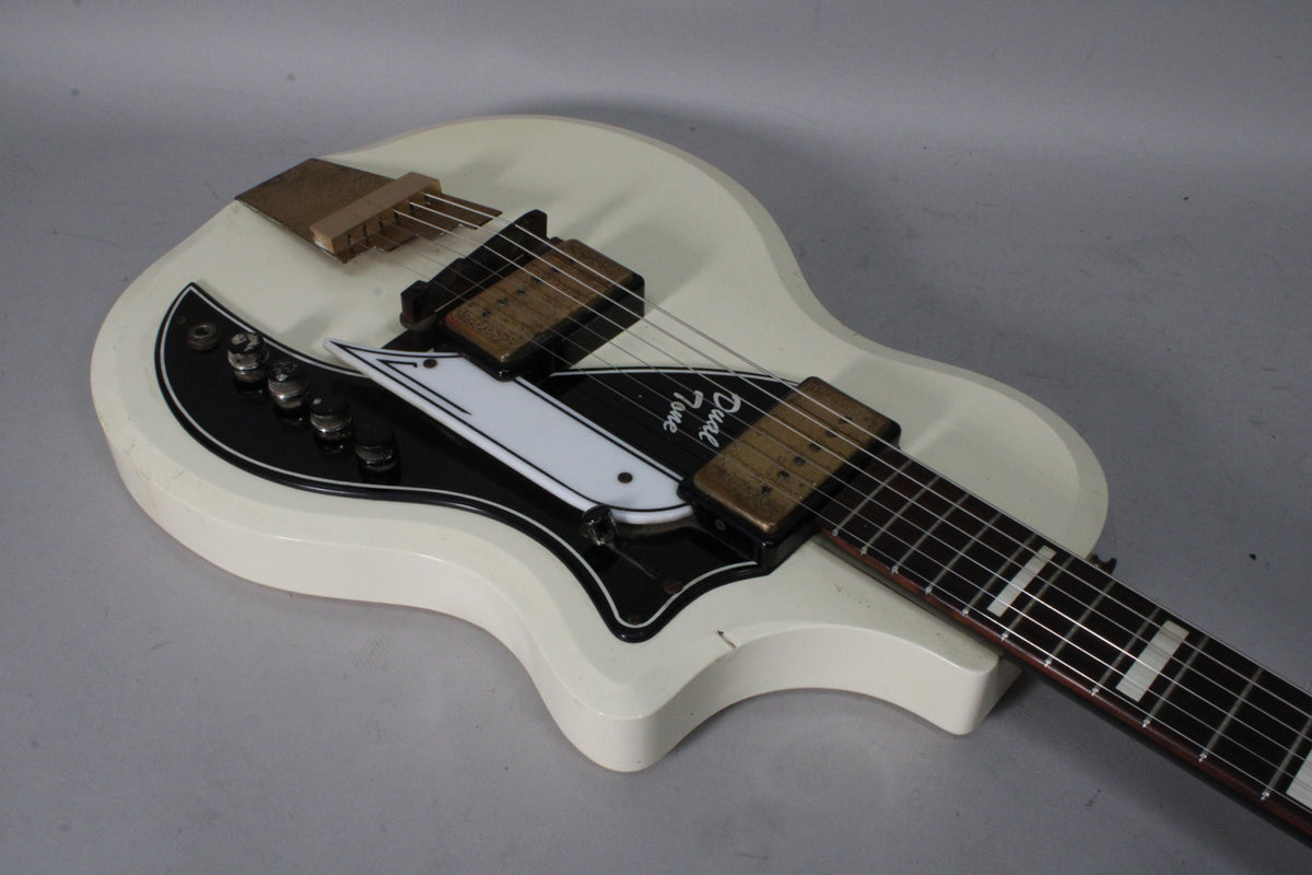The white electric guitar, angled from above, shows its black and white pickguard, four control knobs, and two gold pickups.