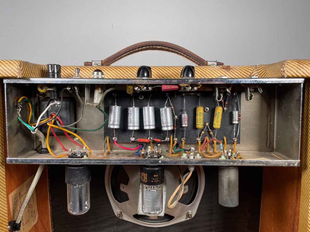 Top-down view of the open tweed amp chassis, revealing internal wiring, capacitors, resistors, and two glowing vacuum tubes.