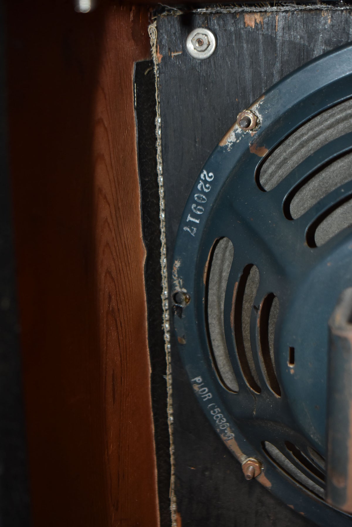 Close-up of a dark blue speaker with visible serial numbers and rust, mounted on worn black wood next to a brown wooden panel.