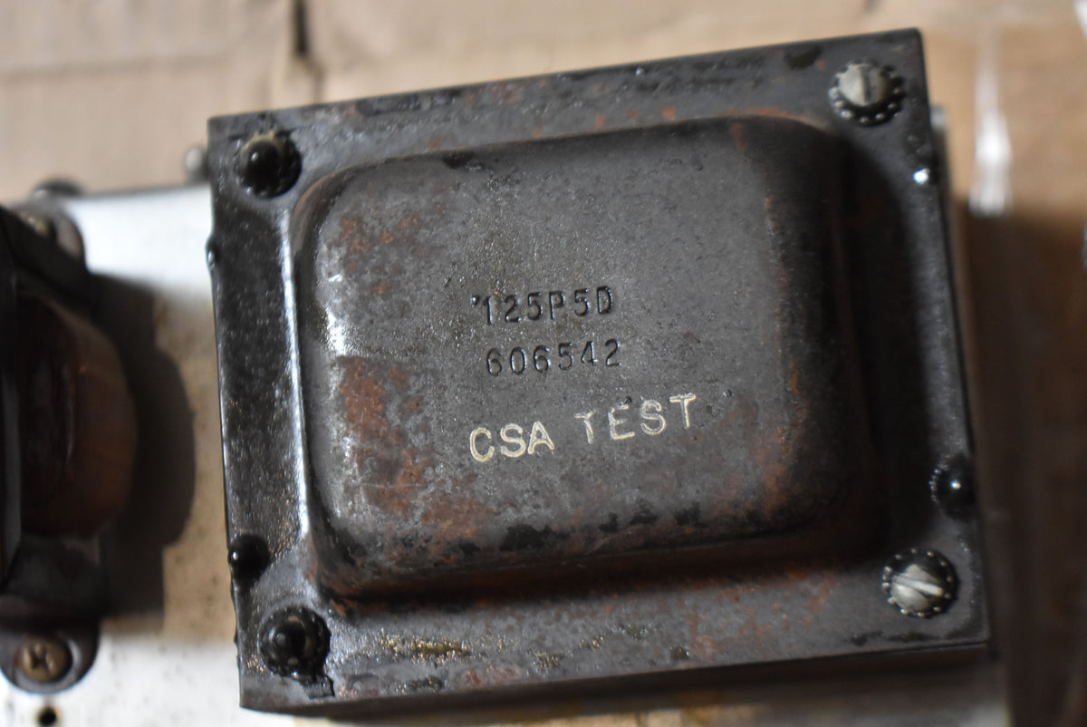 Close-up of a dark, rusty metal transformer with visible numbers and "CSA TEST" text, secured by four screws.