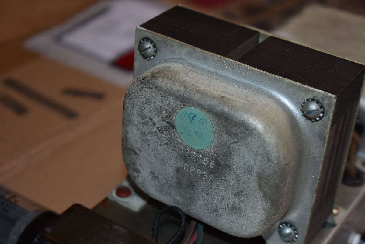 Close-up of a tarnished silver transformer with a faded green sticker and stamped numbers, secured by four screws.