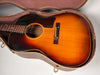 The sunburst acoustic guitar, showing its body, soundhole, pickguard, and bridge, rests in an open, worn brown hard case.
