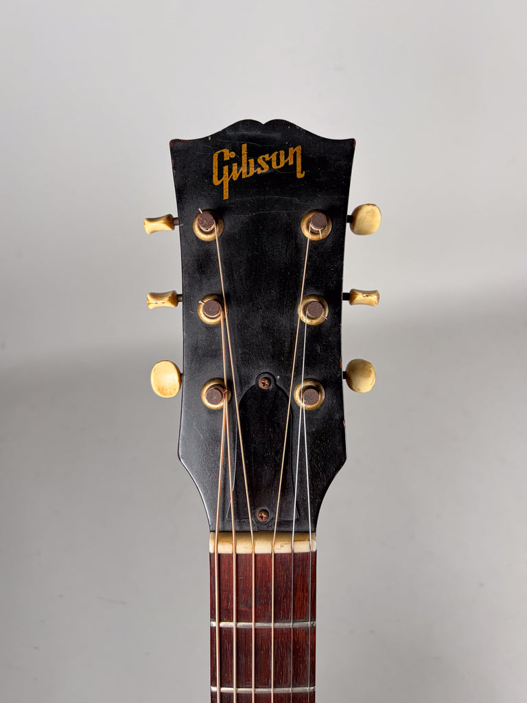 The guitar's black headstock, with gold lettering and six cream tuning pegs, shows significant wear and finish checking.