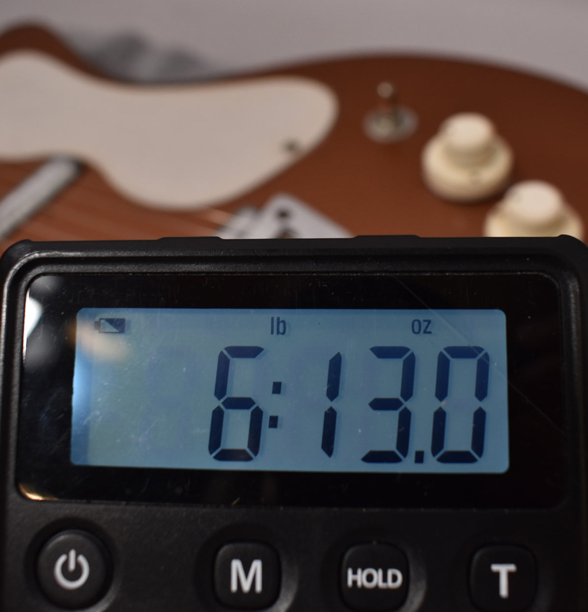 A digital scale displays "6:13.0 lb oz" with the guitar's copper body and cream pickguard blurred in the background.