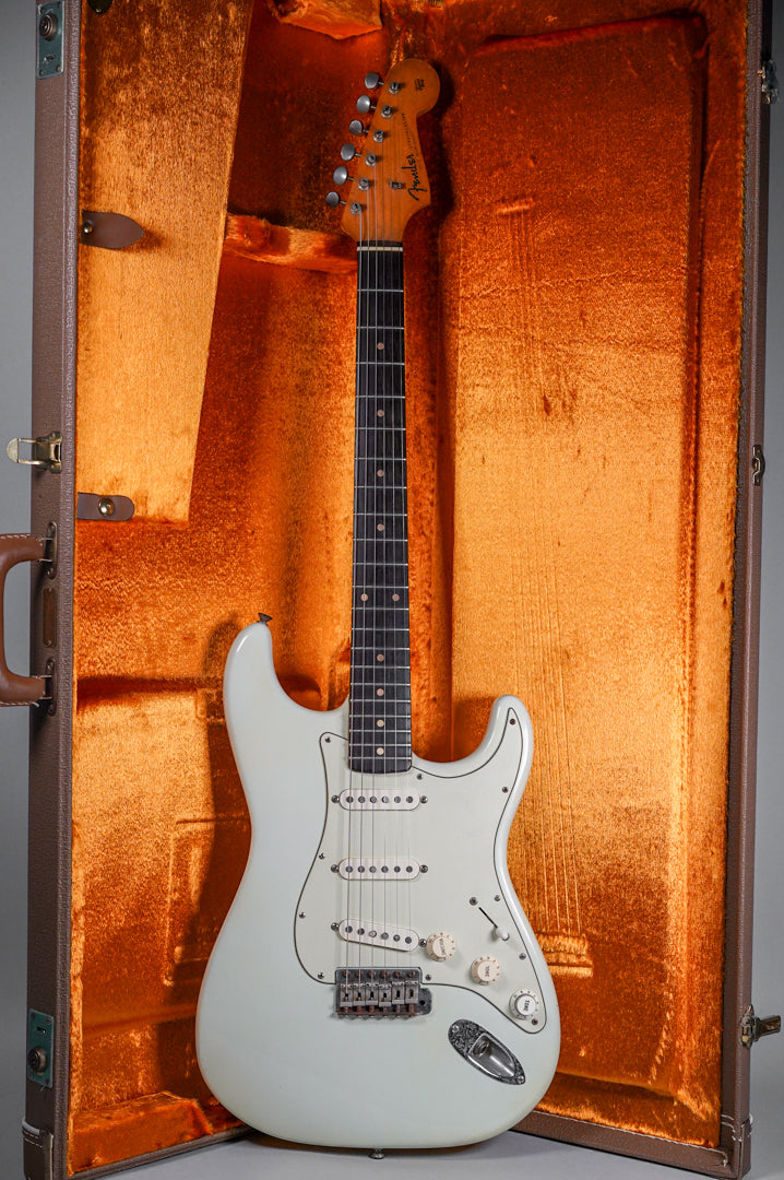 Full view of a white electric guitar with a dark fretboard, three single-coil pickups, and a tremolo bridge, standing in an open brown hardshell case with orange plush lining.