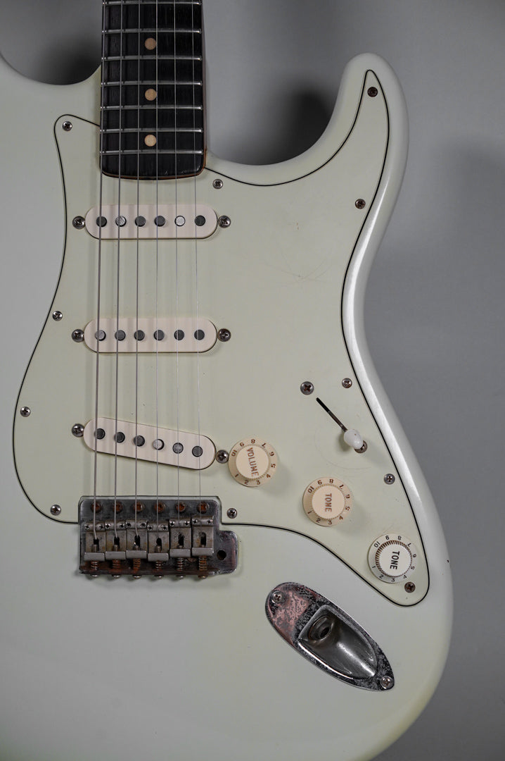 Close-up of a white electric guitar body, showing the fretboard, three single-coil pickups, three cream control knobs, and a rusted tremolo bridge.