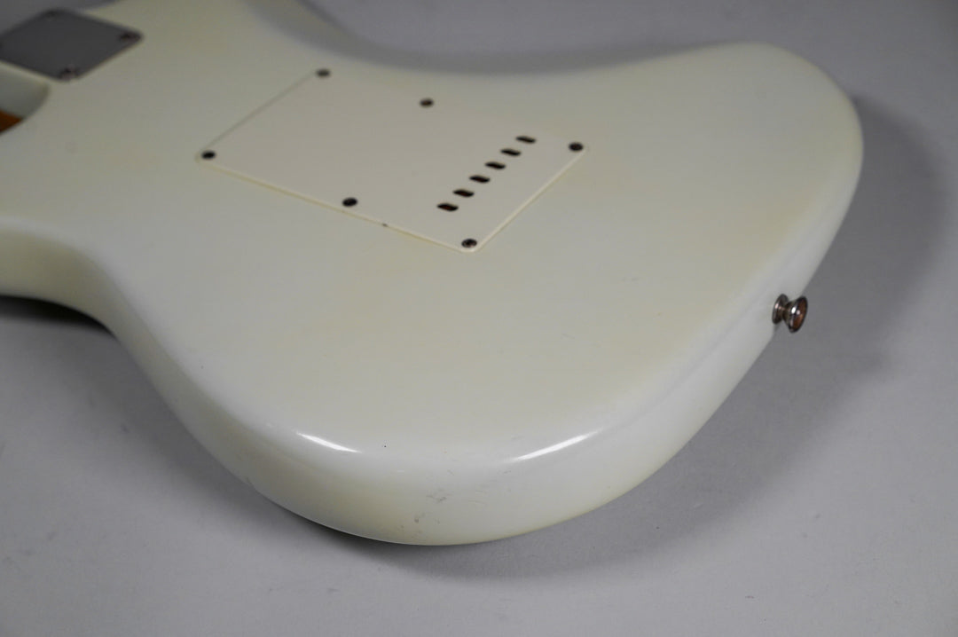 The back of a white electric guitar, angled to show the tremolo cavity cover and a strap button, with minor wear.