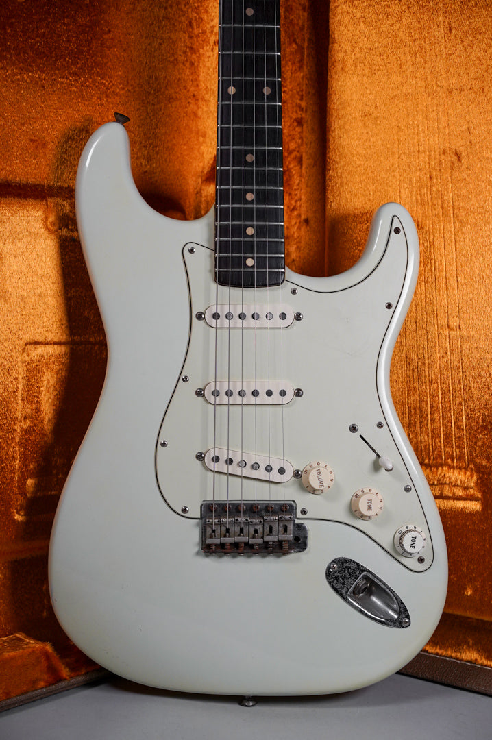 The white electric guitar is angled, showing its body, three single-coil pickups, three white control knobs, and a chrome bridge.