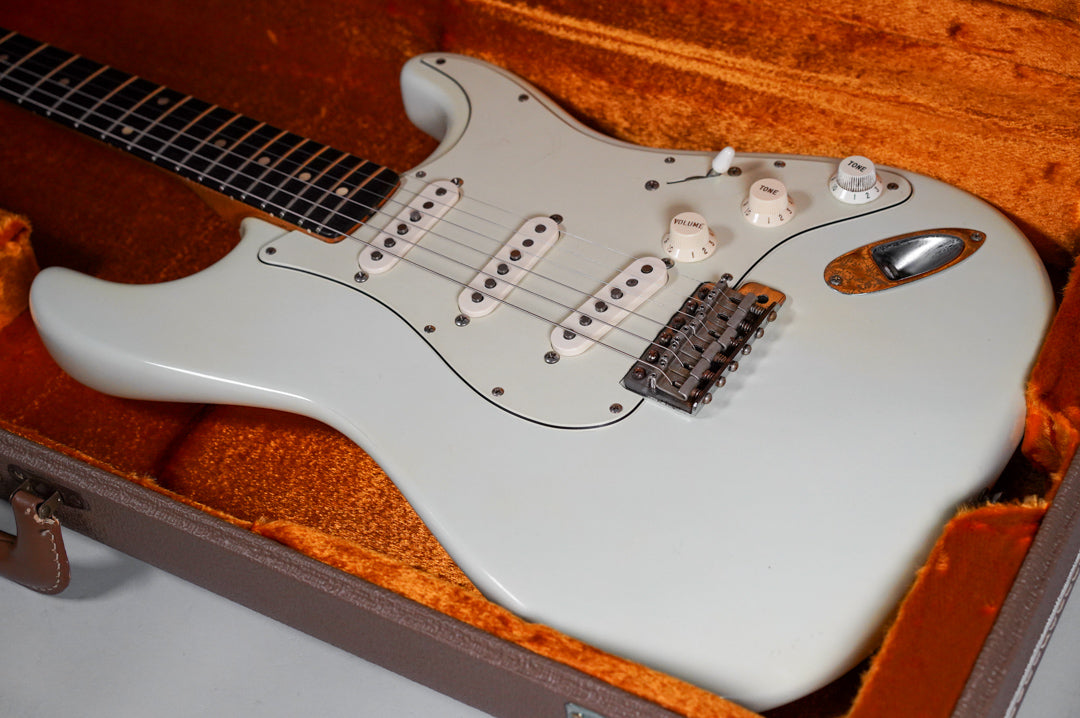 The white refinished electric guitar, with three single-coil pickups and aged white controls, rests in a brown case with orange lining.