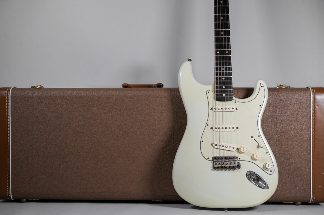 Full view of a white electric guitar with a dark fretboard, standing upright next to a brown hard shell case.