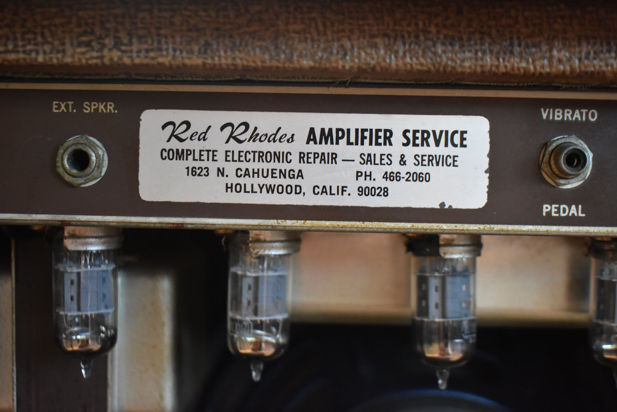 Close-up of the brown control panel showing the "Red Rhodes Amplifier Service" sticker, two input jacks, and four glowing vacuum tubes.