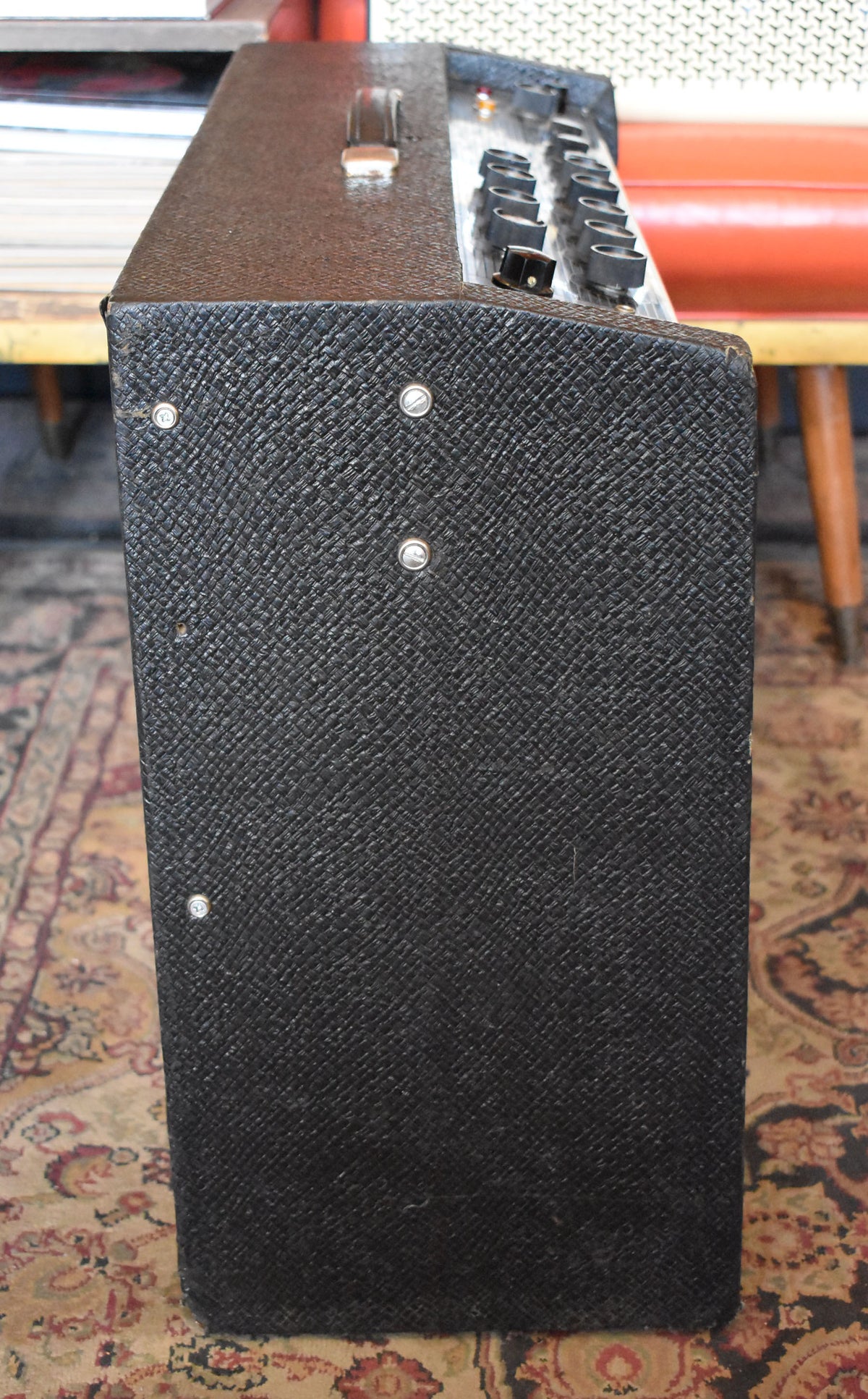The black textured amp is angled right, showing its side with four silver screws and the top control panel with black knobs.