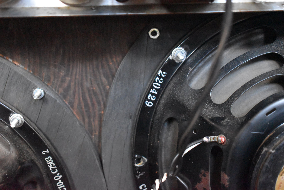Close-up of two dark grey speakers with visible dust, mounted on a dark wood panel, showing wiring and mounting bolts.