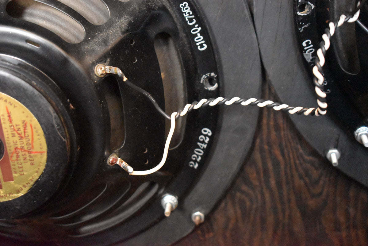Close-up of two black speakers with white and black twisted wires, showing a gold and red label on the left speaker.