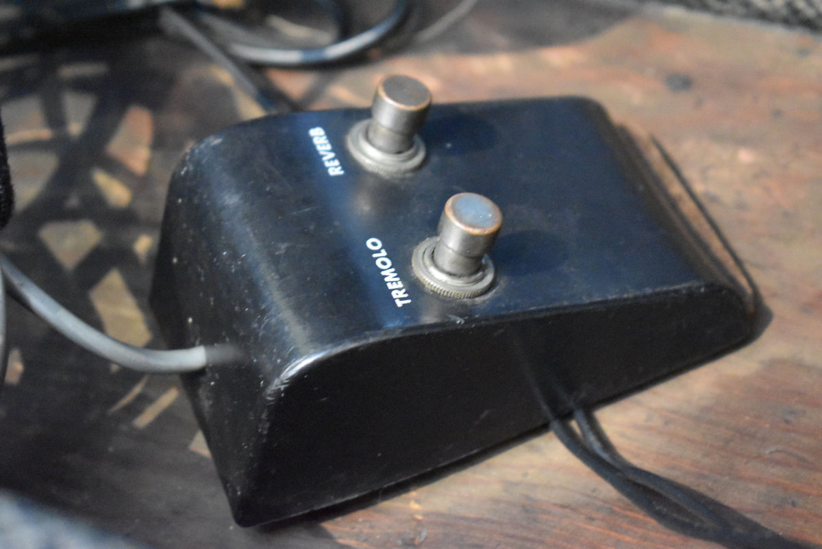 Black two-button footswitch with "REVERB" and "TREMOLO" labels, showing wear on the metal buttons and scuffs on the plastic casing.